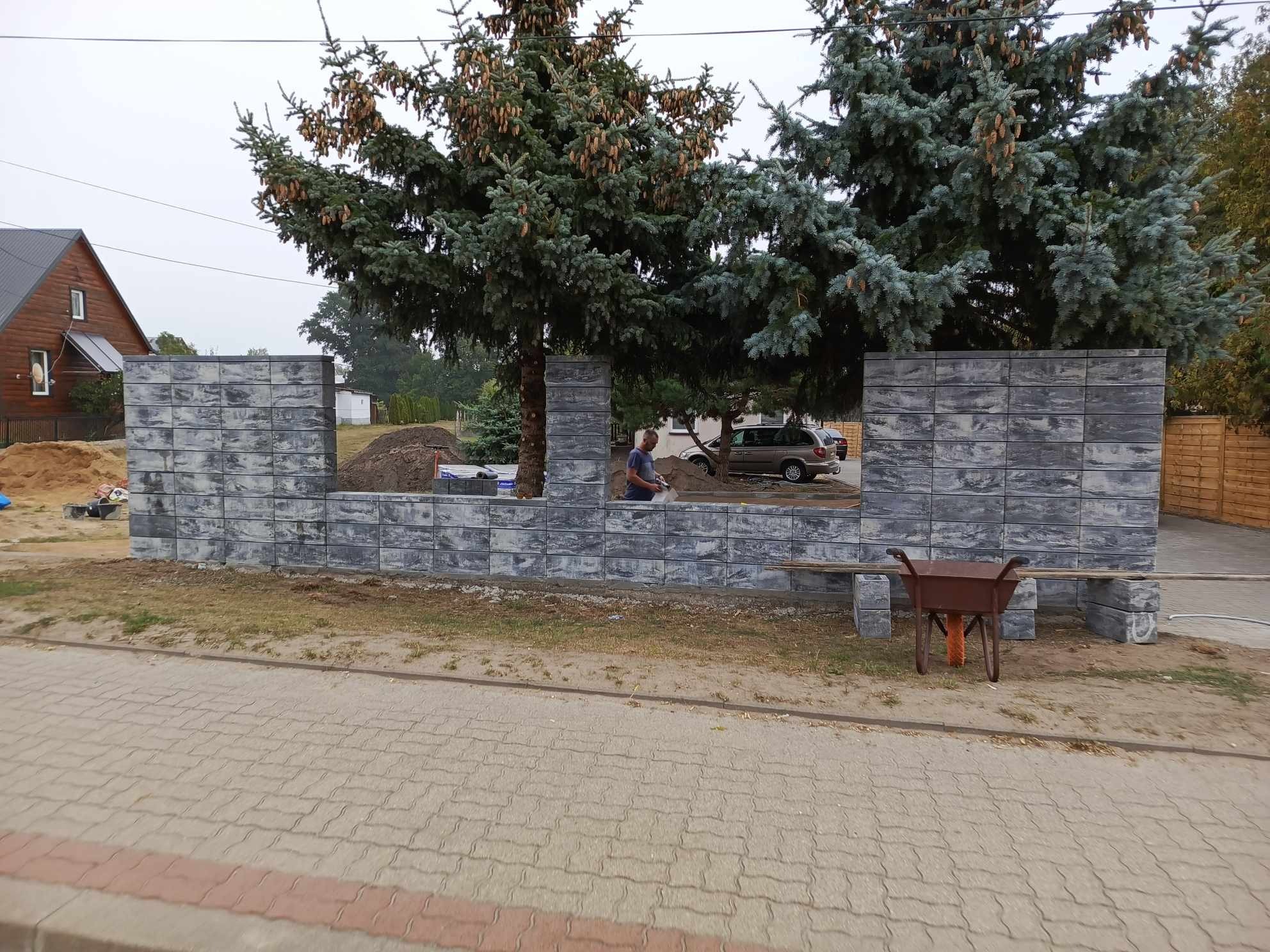 Budowa ogrodzenia z szarych, betonowych bloczków; widoczny murarz przy pracy, taczka i stosy materiałów budowlanych w tle.