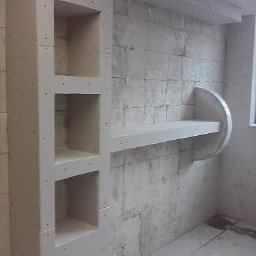 Łukasz Zych - Surowy stan: Ściana z cegły pokryta płytą gipsowo-kartonową z wnękami i półką w trakcie montażu, widoczne ślady szpachlowania i brak wykończenia.