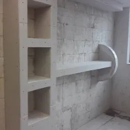 Surowy stan: Ściana z cegły pokryta płytą gipsowo-kartonową z wnękami i półką w trakcie montażu, widoczne ślady szpachlowania i brak wykończenia.