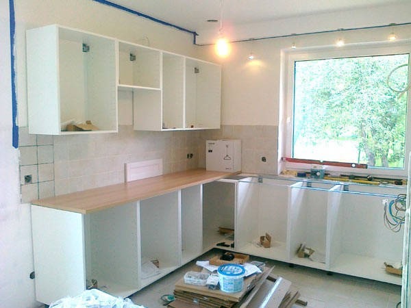 Wnętrze kuchni w trakcie montażu: białe szafki wiszące i stojące bez frontów, drewniany blat, narzędzia i materiały budowlane widoczne na podłodze, okno z widokiem na zieleń.