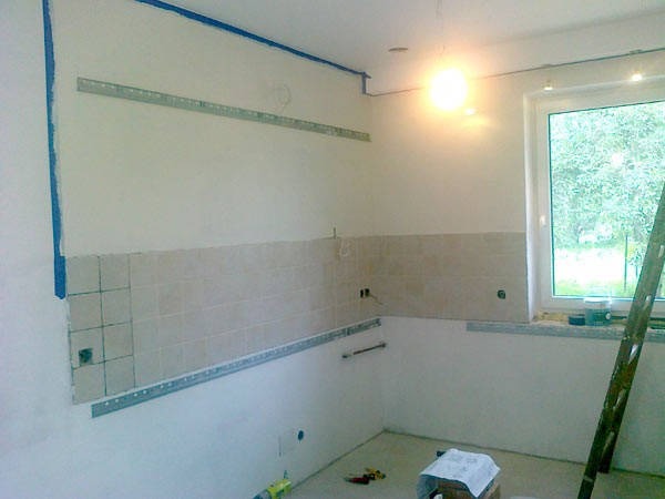 Wnętrze kuchni w trakcie remontu: częściowo położone beżowe płytki, metalowe listwy montażowe przy ścianach, zabezpieczone taśmą malarską narożniki, drabina oparta o okno.