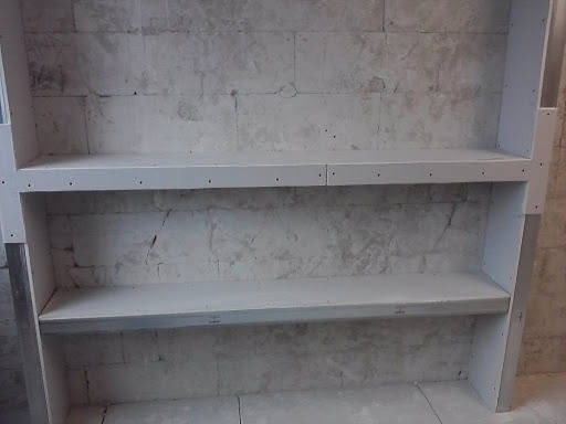 Niedokończony biały regał z dwiema półkami na tle surowej, szarej ściany z cegieł, widoczne ślady po wierceniu.