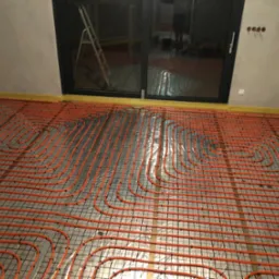 Pomarańczowe rury ogrzewania podłogowego ułożone na czarnej siatce w surowym wnętrzu, widoczne okno balkonowe i drabina.