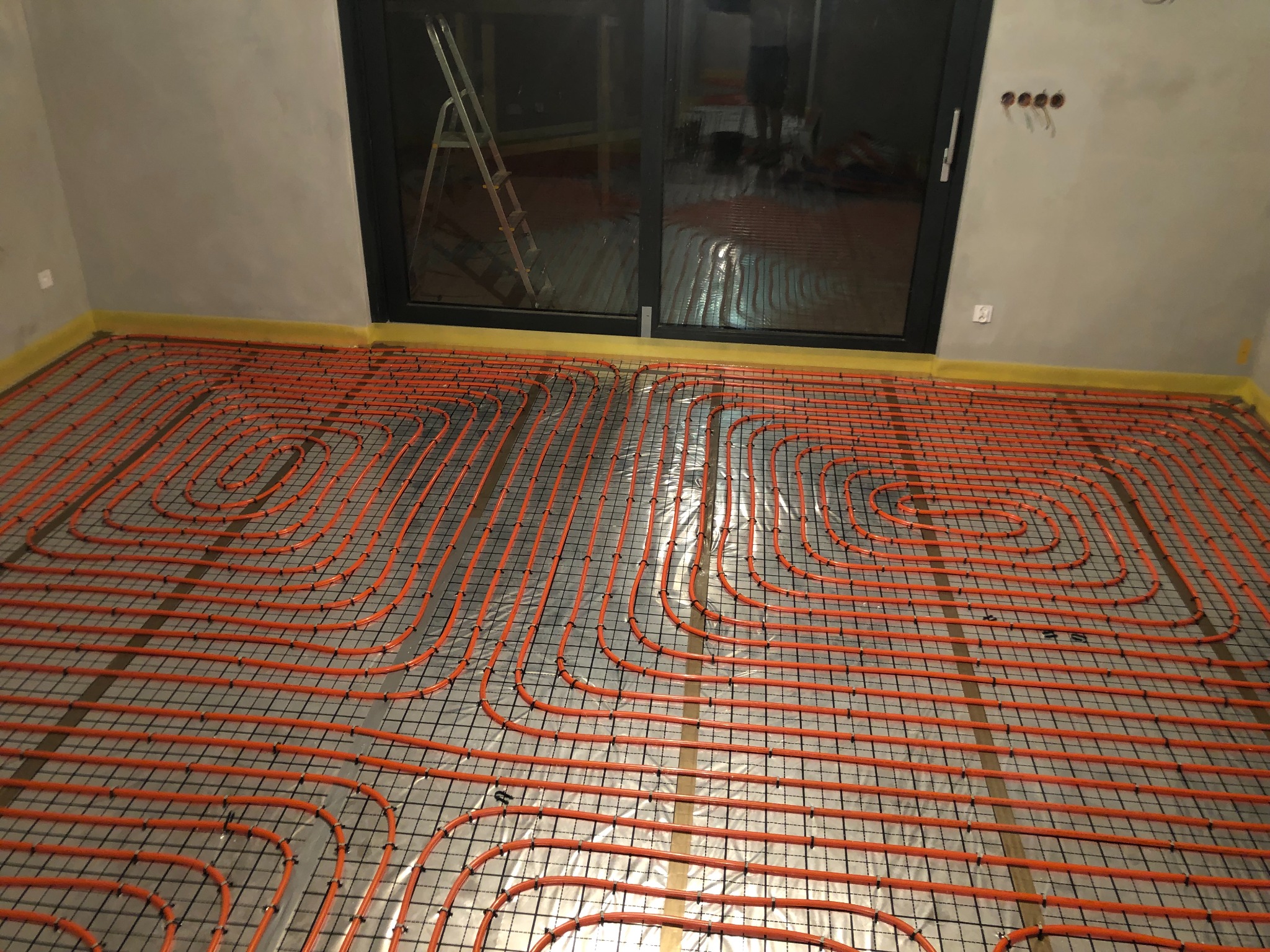 Pomarańczowe rury ogrzewania podłogowego ułożone na czarnej siatce w surowym wnętrzu, widoczne okno balkonowe i drabina.
