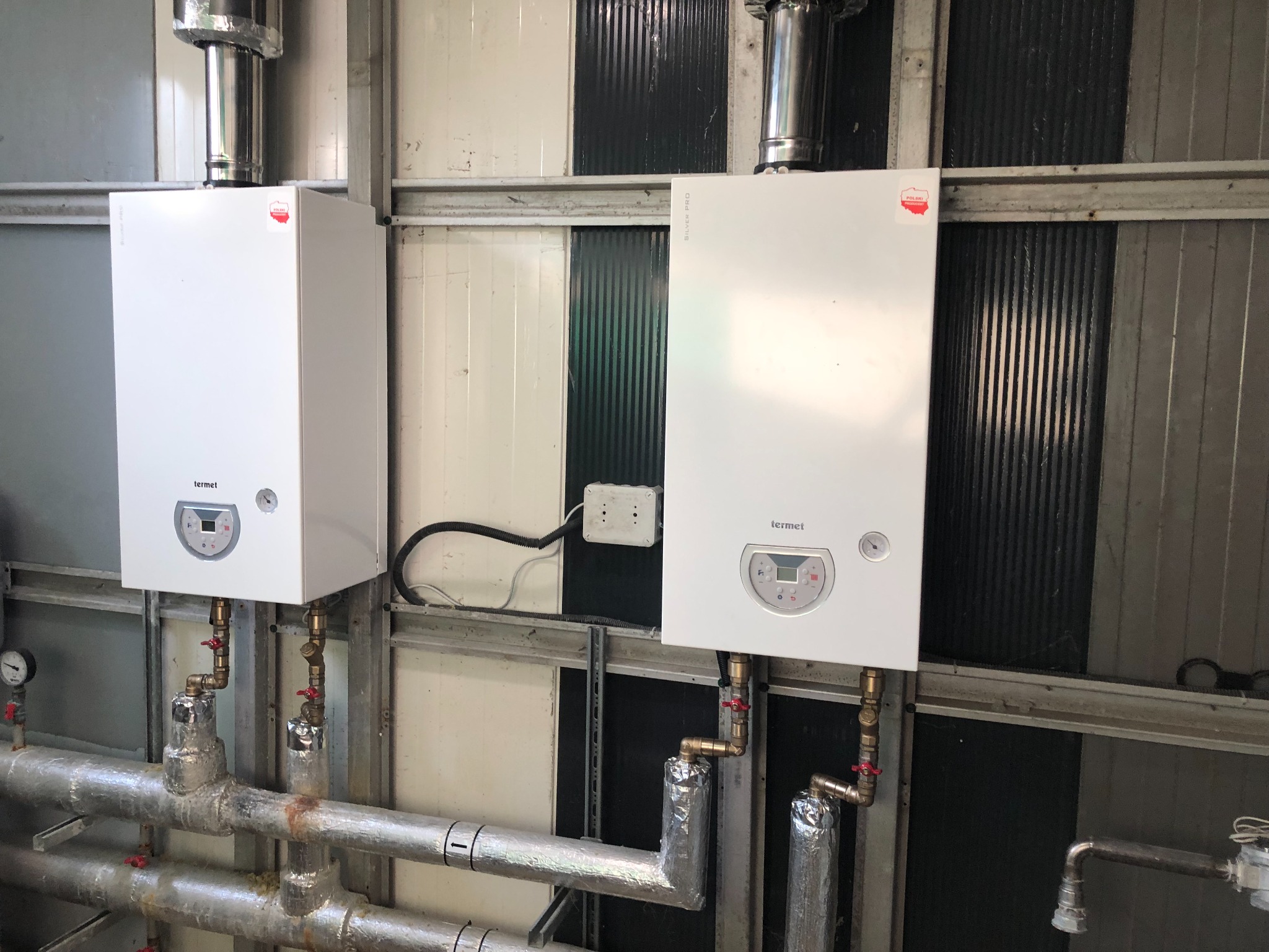 Dwa białe piece grzewcze marki Termet zamontowane na ścianie z widocznymi rurami instalacji, zaworami i izolacją termiczną.