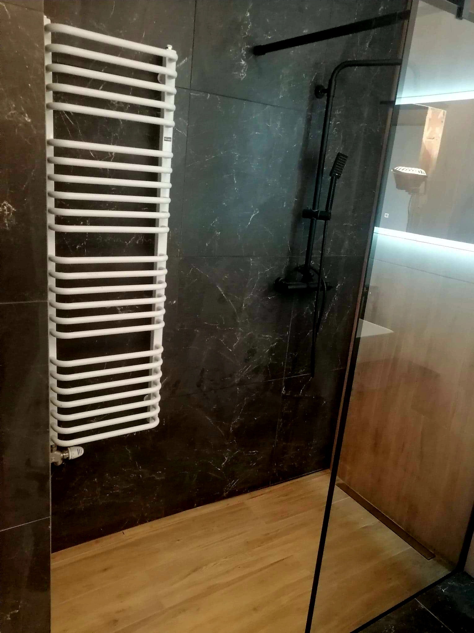 Nowoczesna łazienka z czarnymi płytkami imitującymi marmur, białym grzejnikiem drabinkowym, czarną armaturą prysznicową i drewnopodobną podłogą.