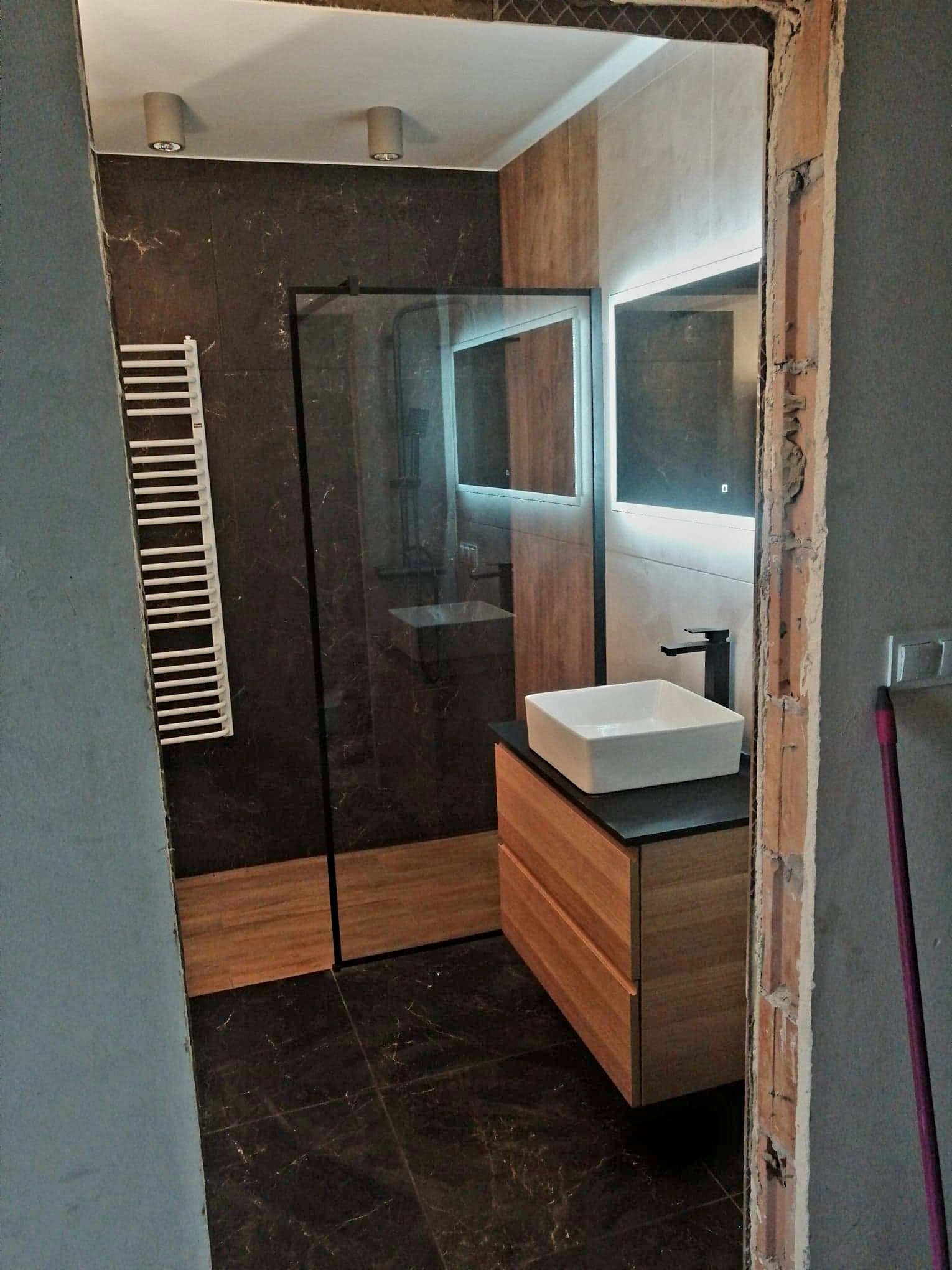 Nowoczesna łazienka z kabiną prysznicową typu walk-in, wiszącą szafką z umywalką nablatową i grzejnikiem drabinkowym, widoczna przez otwór w ścianie w trakcie prac wykończeniowych.