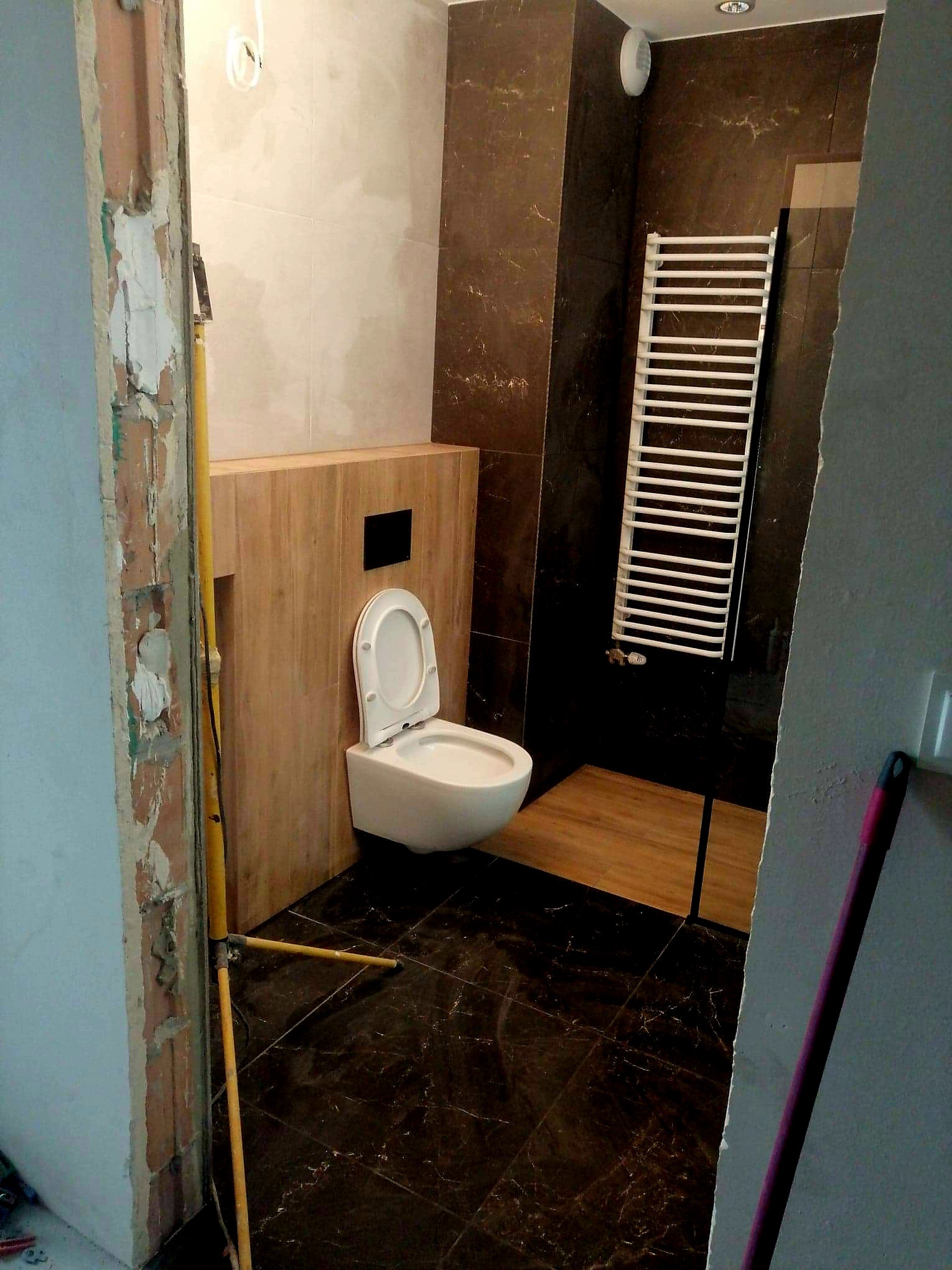 Remont łazienki w trakcie: wisząca toaleta z otwartą deską, brązowe marmurowe płytki podłogowe i ścienne, biały grzejnik drabinkowy, surowe wykończenie ościeżnicy.