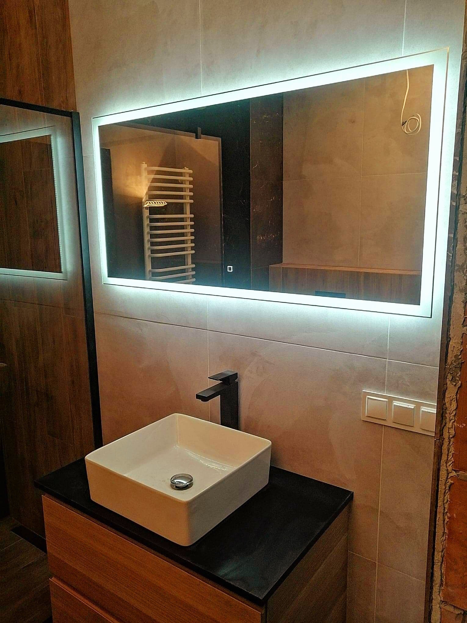 Nowoczesna łazienka z podświetlanym lustrem, kwadratową umywalką nablatową i czarną armaturą. Fragment ściany z widoczną surową cegłą w tle.