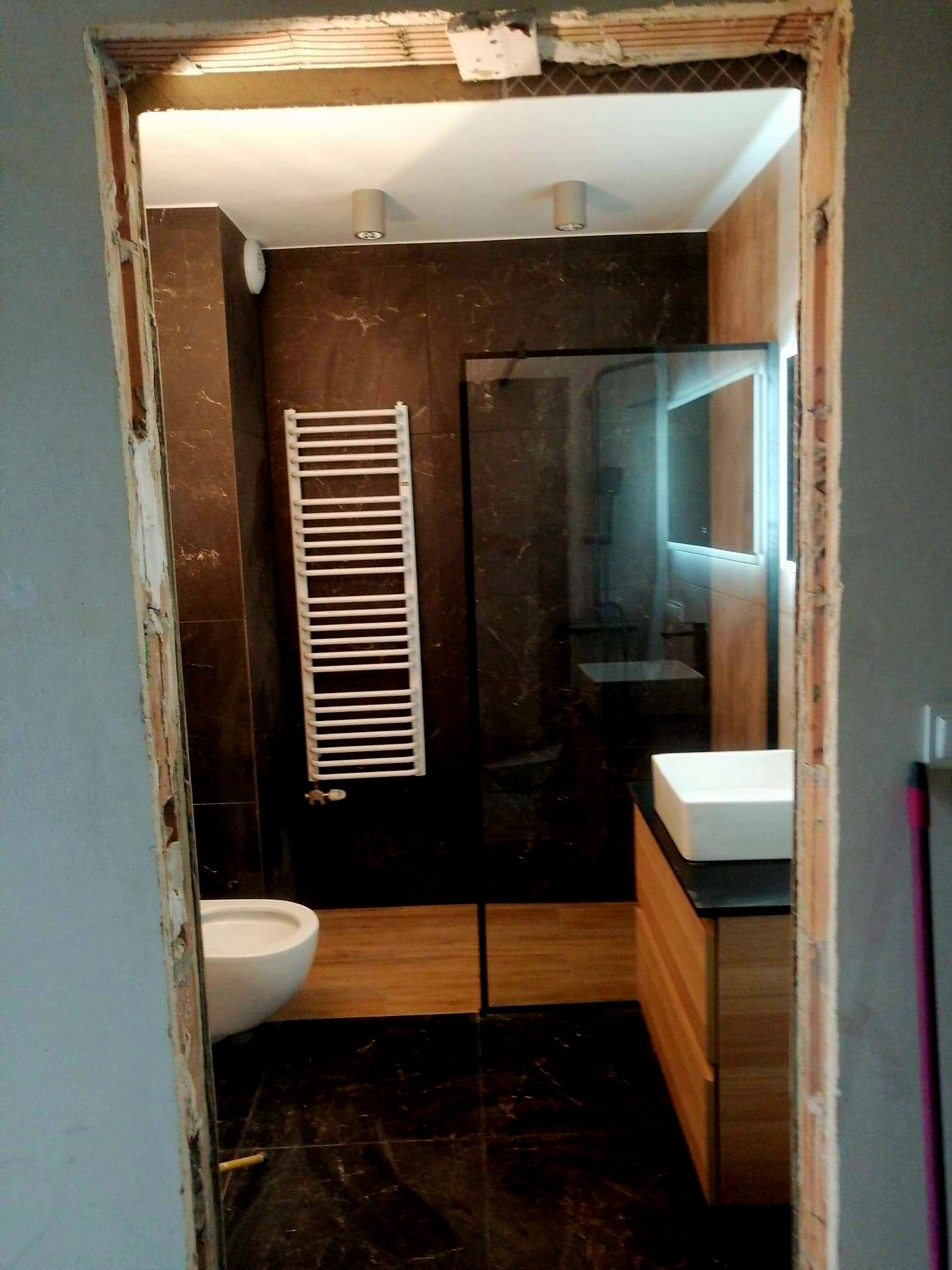 Wnętrze łazienki w trakcie prac, widok przez surową futrynę, widoczne płytki imitujące marmur, grzejnik drabinkowy, toaleta wisząca i umywalka nablatowa.