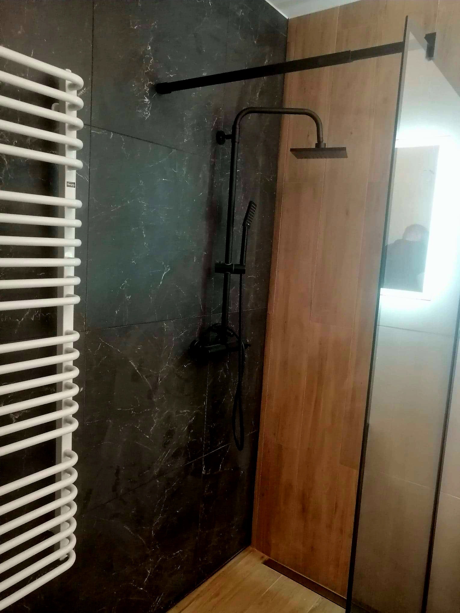 Nowoczesna łazienka z czarnymi płytkami imitującymi marmur, drewnianą ścianką prysznicową i czarną armaturą, w rogu widoczny biały grzejnik drabinkowy.