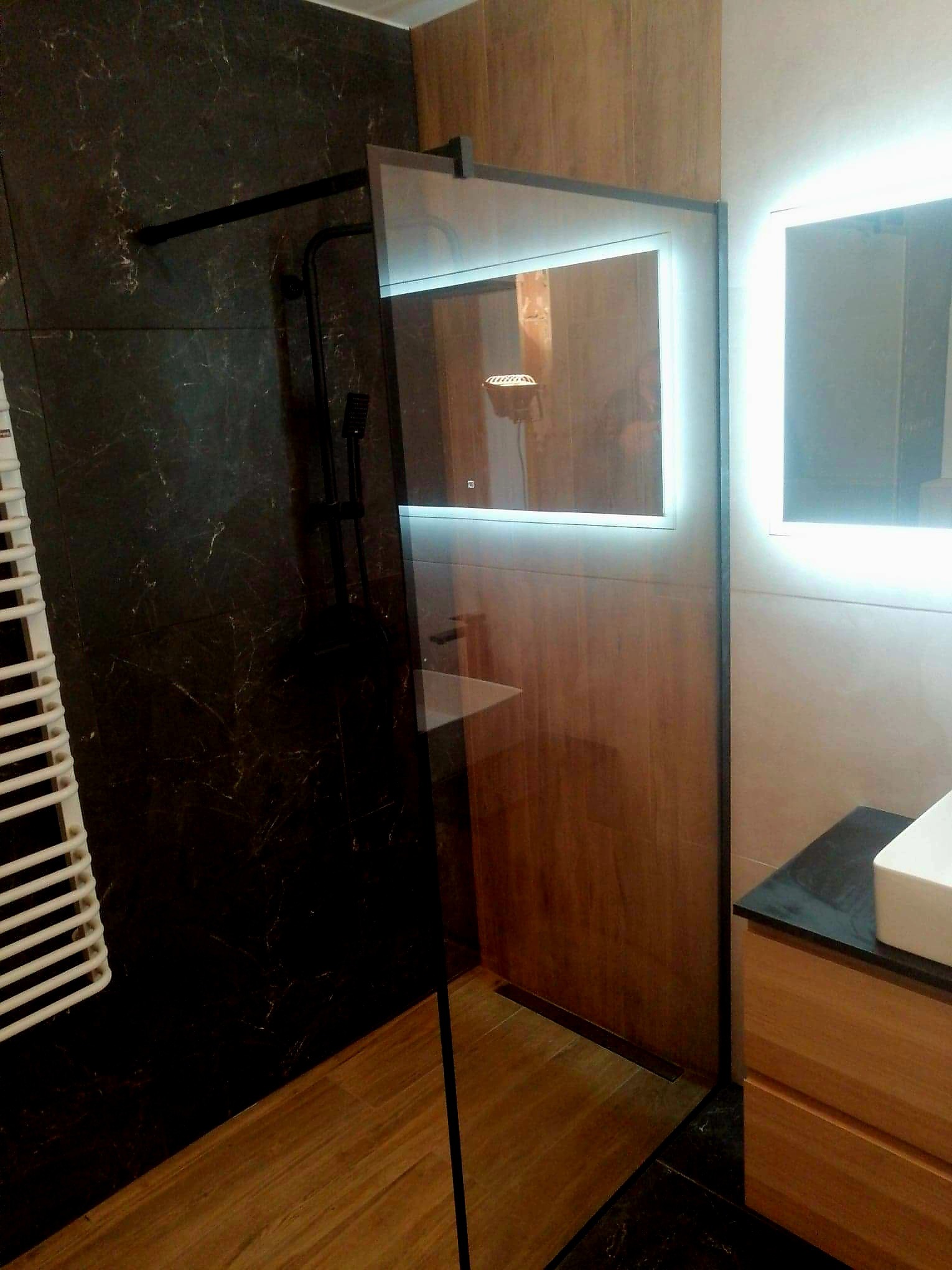 Nowoczesna łazienka z czarnymi płytkami imitującymi marmur, drewnianymi panelami, szklaną kabiną prysznicową z czarnymi profilami, podświetlanym lustrem i drewnianą szafką pod umywalką.