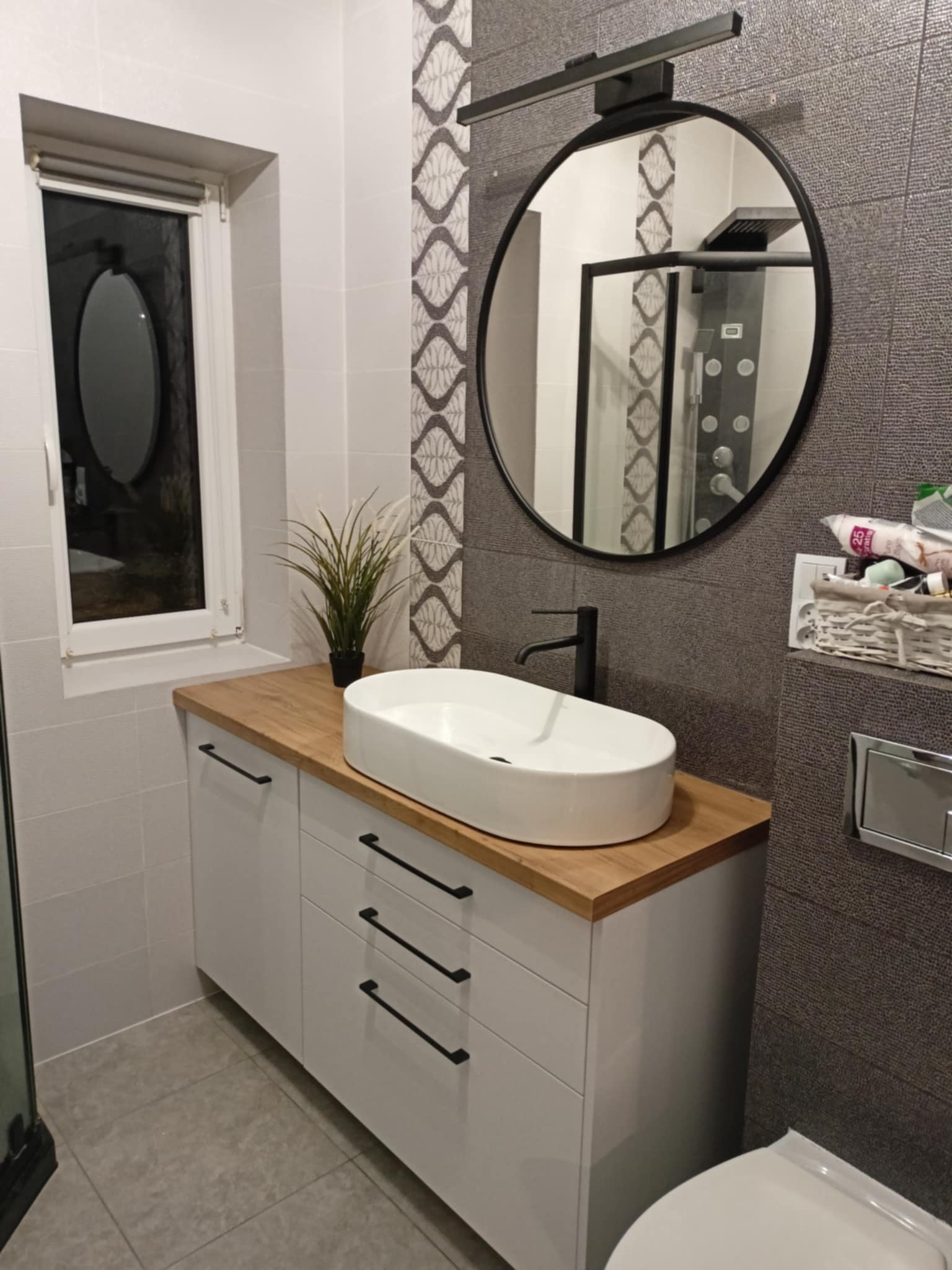 Nowoczesna łazienka z owalną umywalką nablatową, czarną armaturą i okrągłym lustrem z podświetleniem. Dekoracyjne płytki w geometryczny wzór. Widoczna kabina prysznicowa w tle.