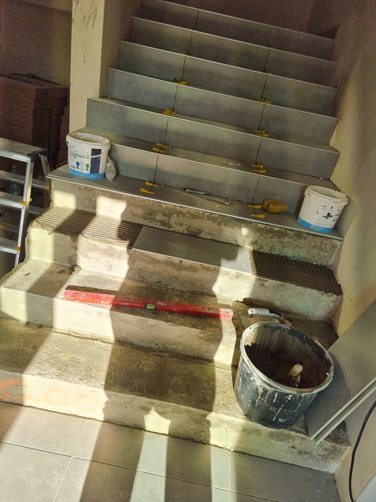 Montaż płytek na schodach z użyciem poziomicy, wiadra z zaprawą i plastikowych krzyżyków dystansowych, widoczne przygotowane podłoże i ułożone płytki.