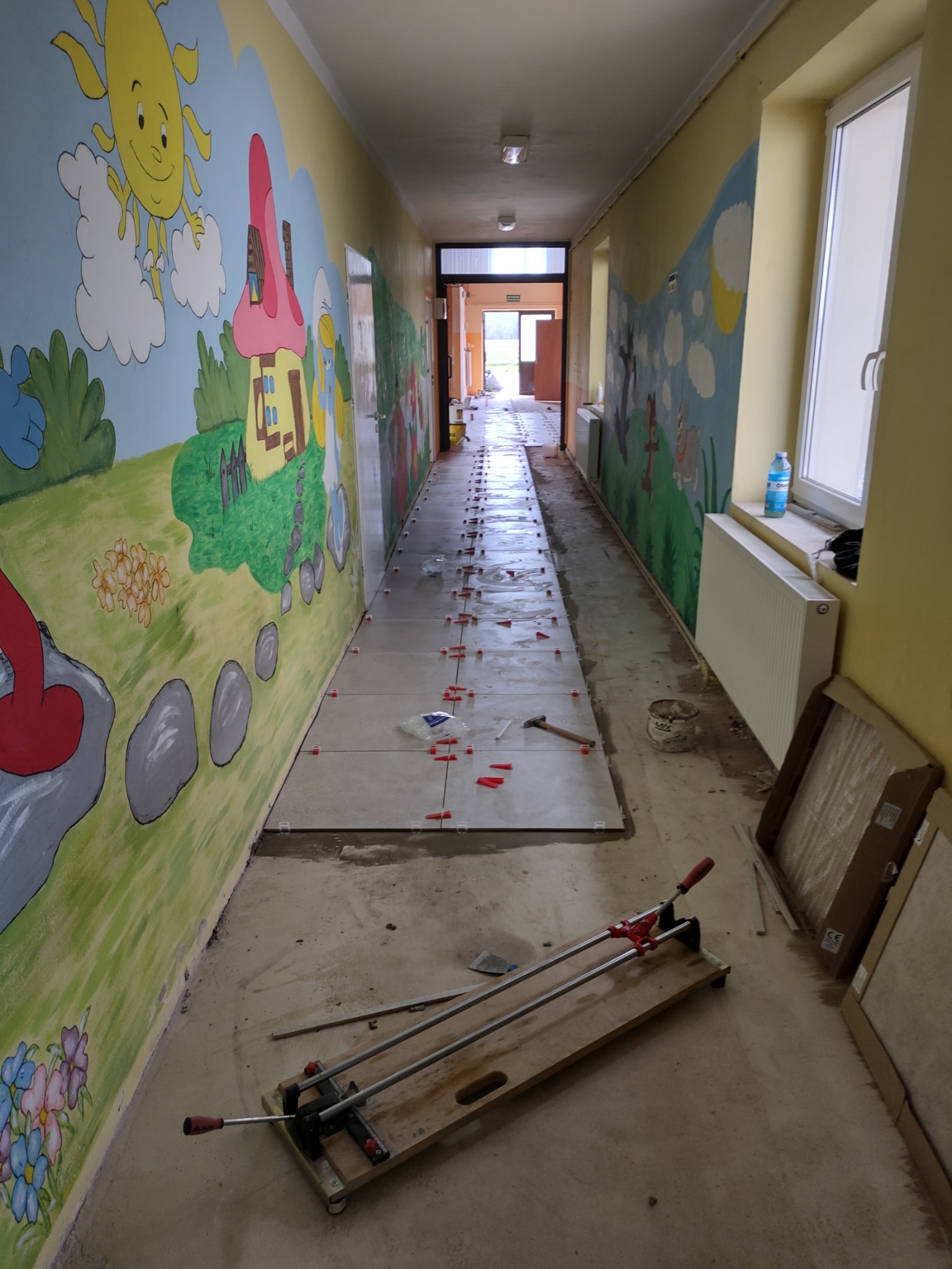 Korytarz w trakcie układania jasnych płytek podłogowych z czerwonymi krzyżykami dystansowymi, na ścianach dziecięce malunki, widoczna przecinarka do płytek i kartony z płytkami.