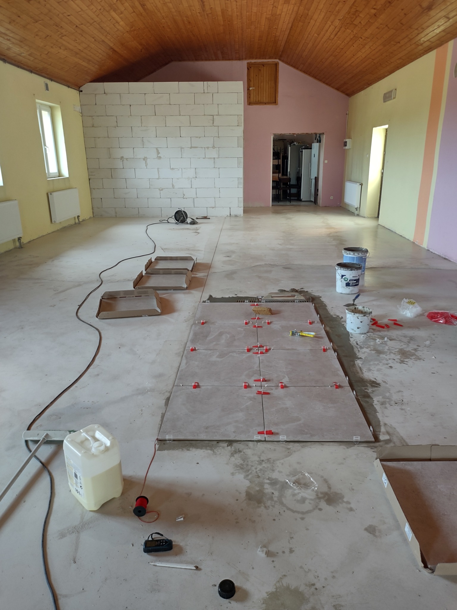 Wnętrze w trakcie prac: częściowo ułożone jasne płytki podłogowe z czerwonymi krzyżykami dystansowymi, widoczne narzędzia, puste wiadra po zaprawie, kartony po płytkach, ściany w surowym stanie...