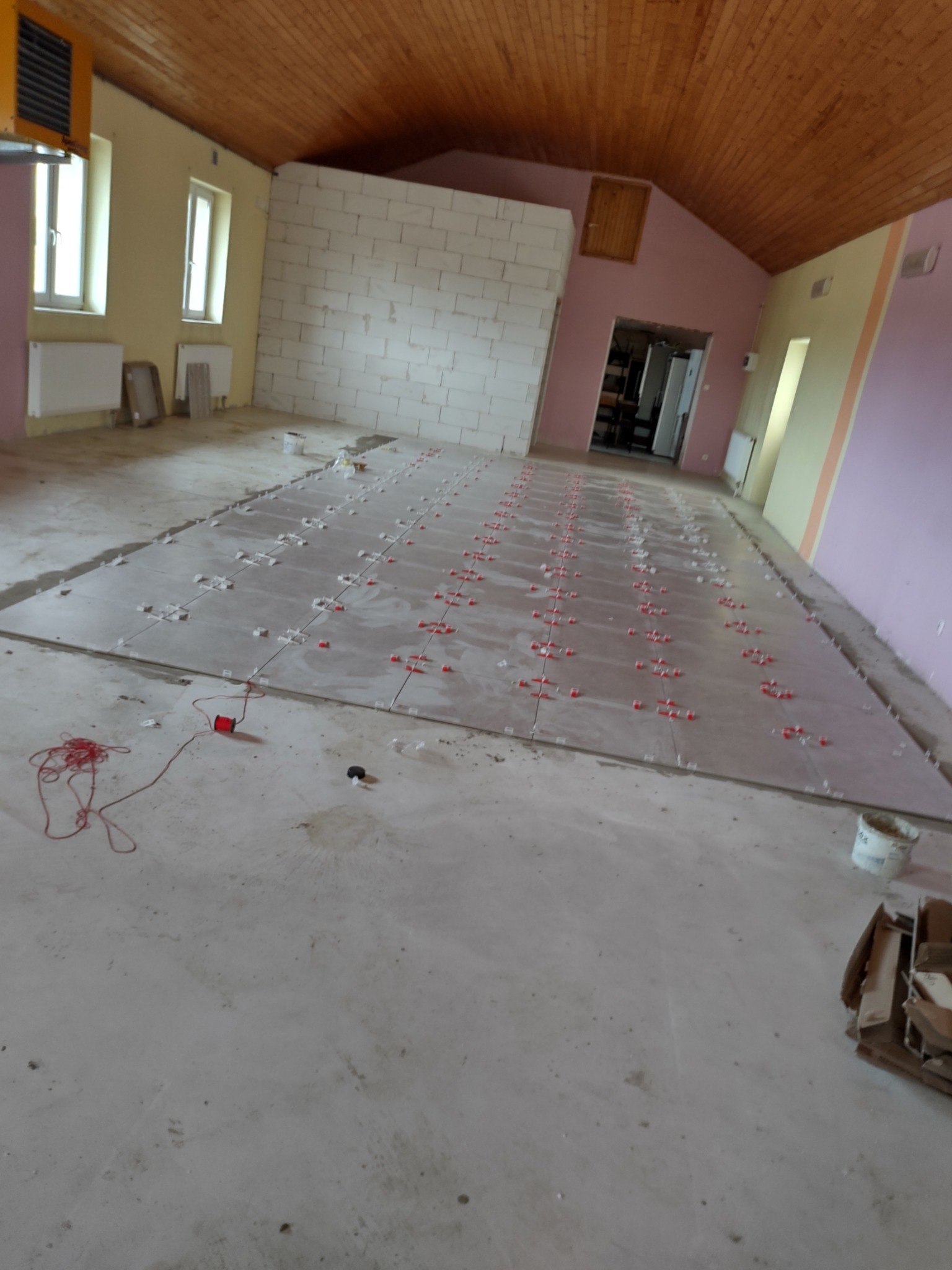 Układanie płytek podłogowych w dużym, surowym pomieszczeniu z drewnianym sufitem, widoczne krzyżyki dystansowe i sznurek traserski.