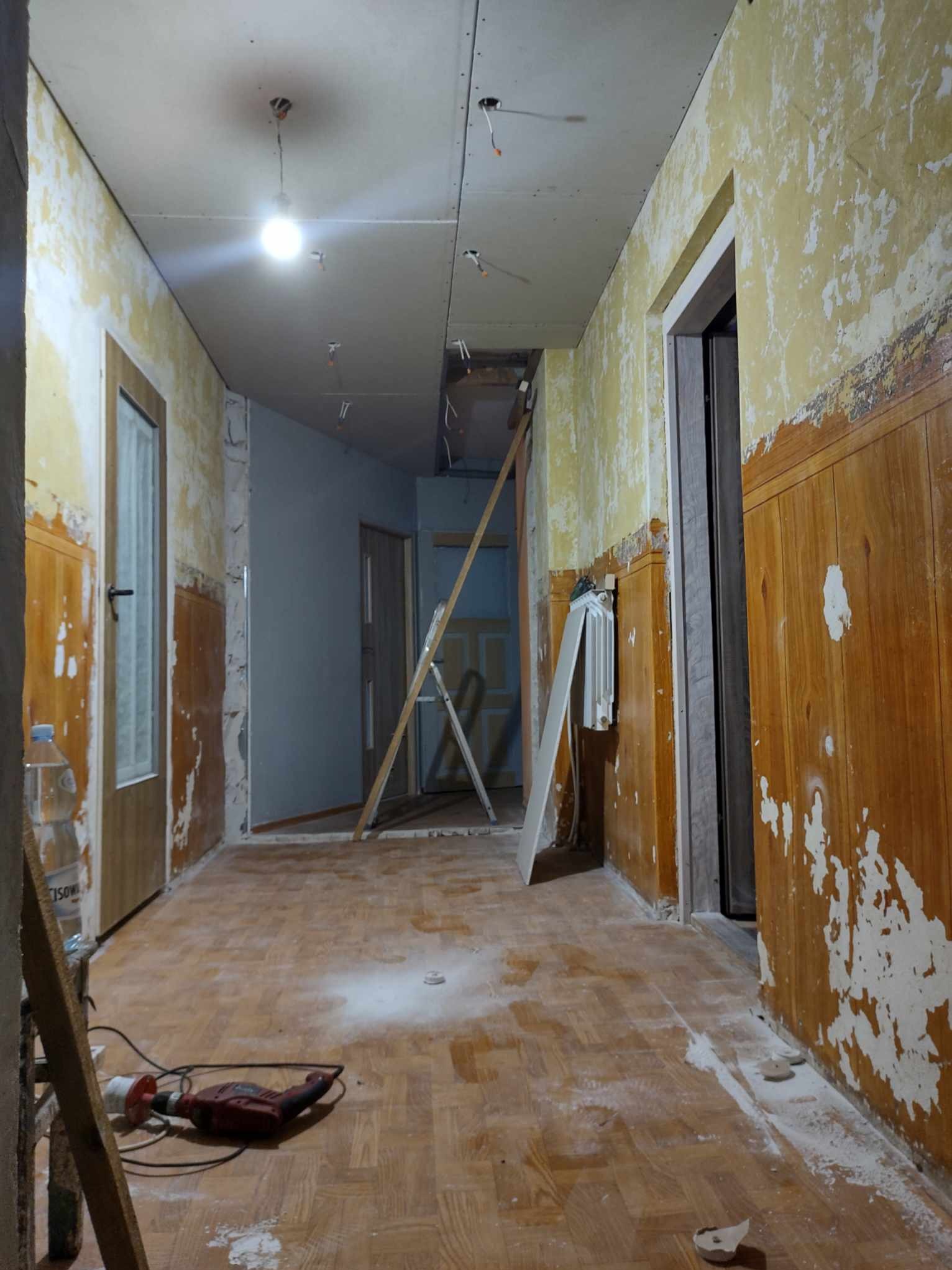 Korytarz w trakcie remontu: zerwana tapeta, płyty karton-gips na suficie, drabina oparta o ścianę, widoczny parkiet i narzędzia pracy, w tym wiertarka.