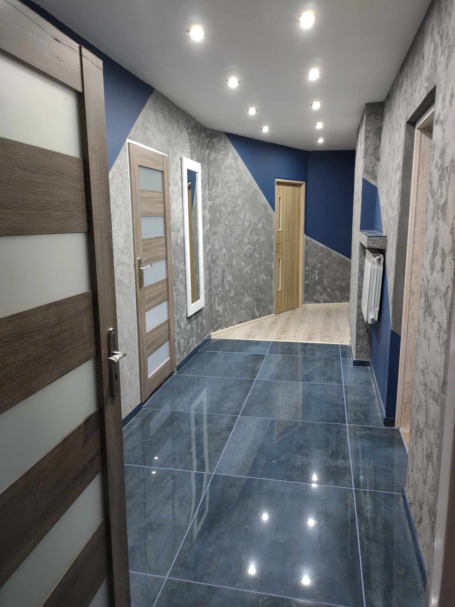 Korytarz po remoncie z ciemnymi płytkami podłogowymi, ścianami w odcieniach szarości i granatu, z widocznym grzejnikiem i kilkoma parami drzwi.
