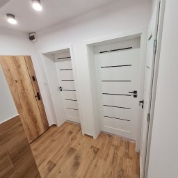 USŁUGI REMONTOWO-WYKOŃCZENIOWE MIKOŁAJ ŁUKOWIAK - Korytarz z trzema drzwiami: jedne drewniane z czarną klamką, dwoje białych z czarnymi, poziomymi paskami i czarnymi klamkami, podłoga wyłożona płytkami imitującymi drewno, oświetlenie sufitowe...
