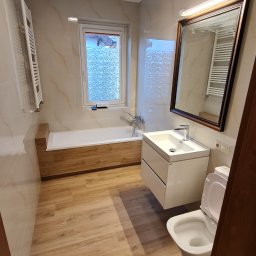 USŁUGI REMONTOWO-WYKOŃCZENIOWE MIKOŁAJ ŁUKOWIAK - Jasna łazienka po remoncie z drewnianą obudową wanny, beżowymi płytkami imitującymi marmur i drewnopodobną podłogą, widok na umywalkę z szafką i toaletę.