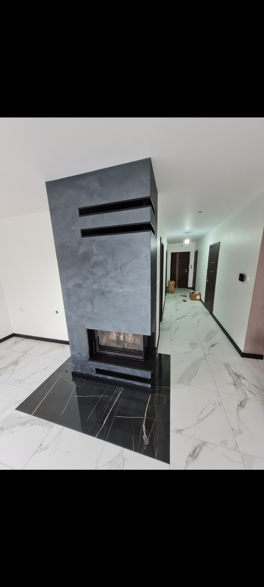 Nowoczesny kominek z szarą ścianą o fakturze betonu i czarnymi wnękami, połączony z podłogą z czarnych płyt marmurowych z białym żyłowaniem, przechodzącą w białą marmurową posadzkę korytarza...