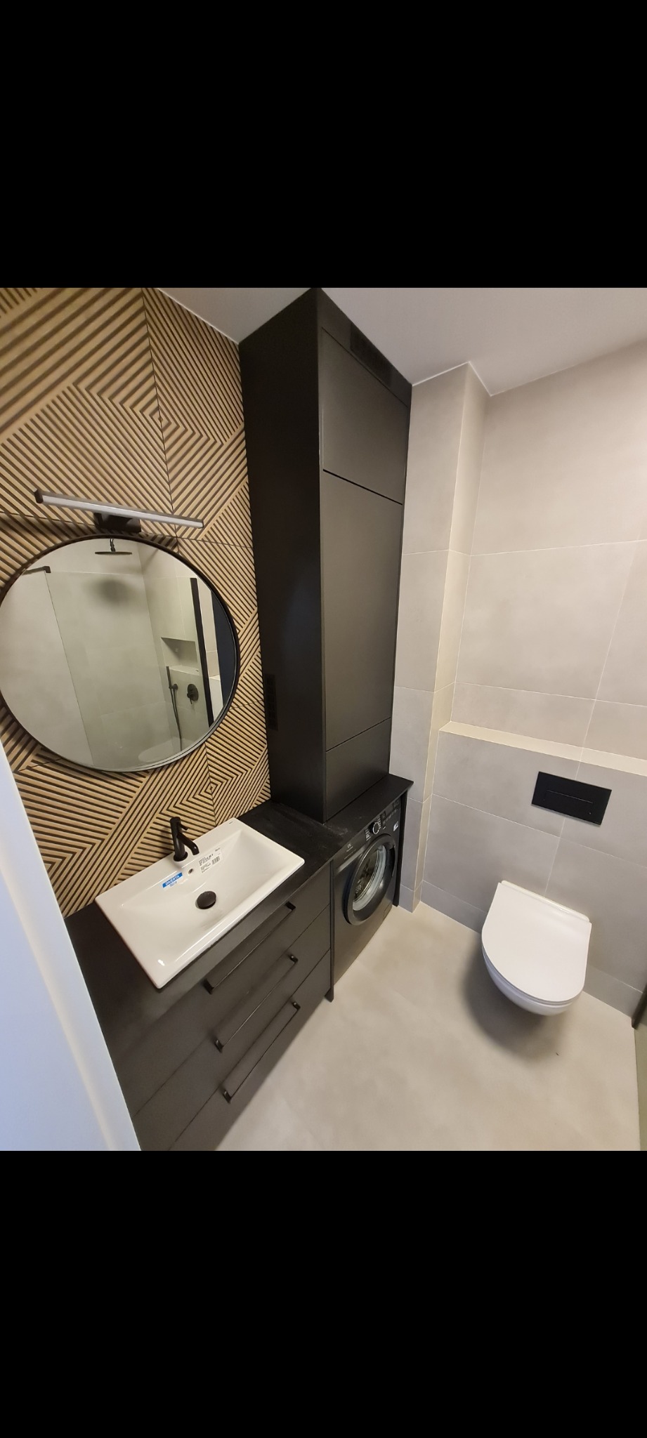 Nowoczesna łazienka z drewnianą ścianą w geometryczne wzory, okrągłym lustrem podświetlanym listwą LED, umywalką nablatową z czarną armaturą, szafką pod umywalką z szufladami, pralką obok szafki...