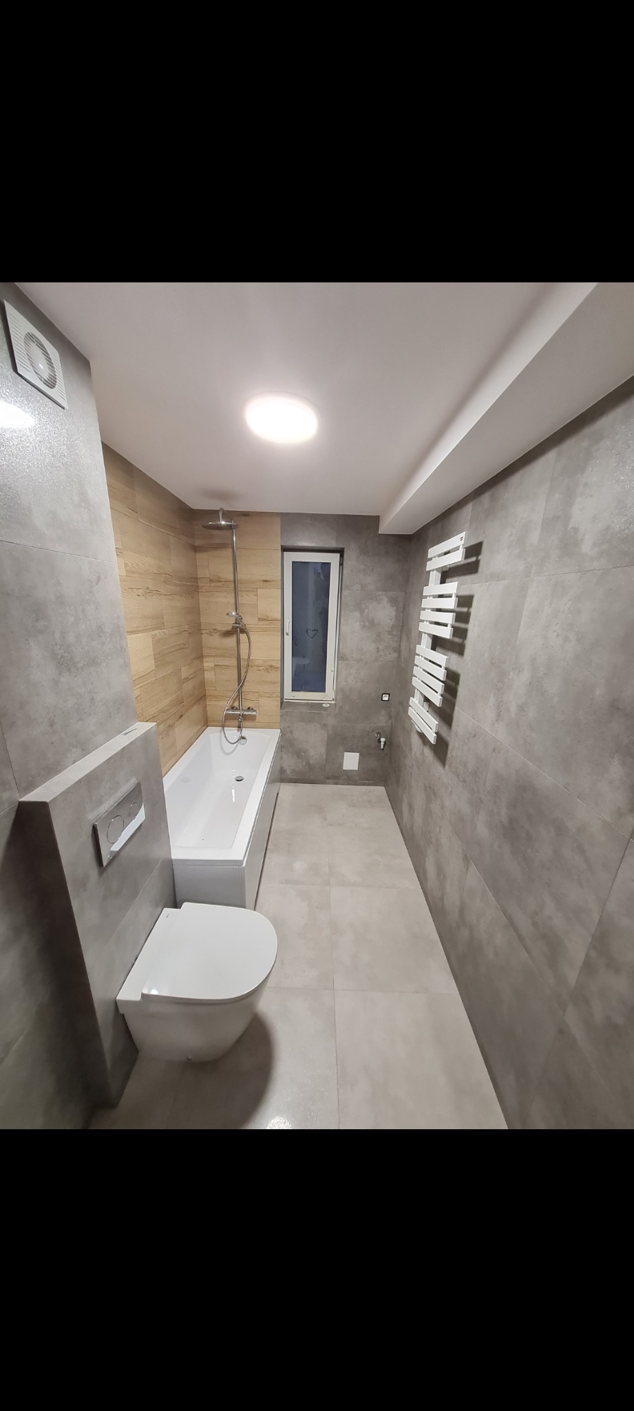 Nowoczesna łazienka po remoncie: szare płytki imitujące beton, drewnopodobna ściana przy wannie, biała wanna i toaleta typu Geberit, grzejnik drabinkowy, małe okno z sercem narysowanym na szybie.