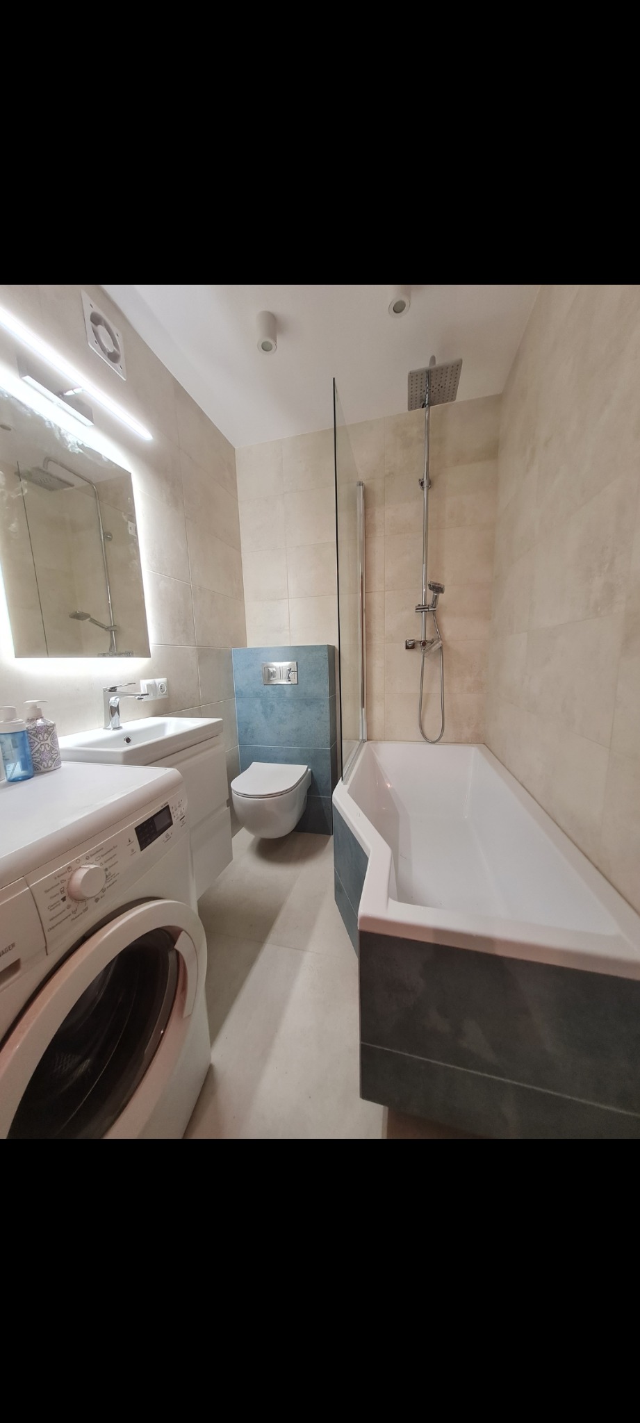 Ukończona łazienka z wanną obudowaną szarą mozaiką, umywalką z szafką, podświetlanym lustrem, toaletą i pralką w jasnej tonacji beżowo-szarej.