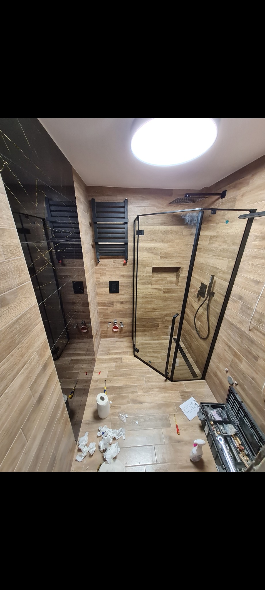 Ukończona łazienka z kabiną prysznicową typu walk-in, czarną armaturą i grzejnikiem drabinkowym, widoczne narzędzia i akcesoria montażowe na podłodze, czarne płytki imitujące marmur na jednej ze...