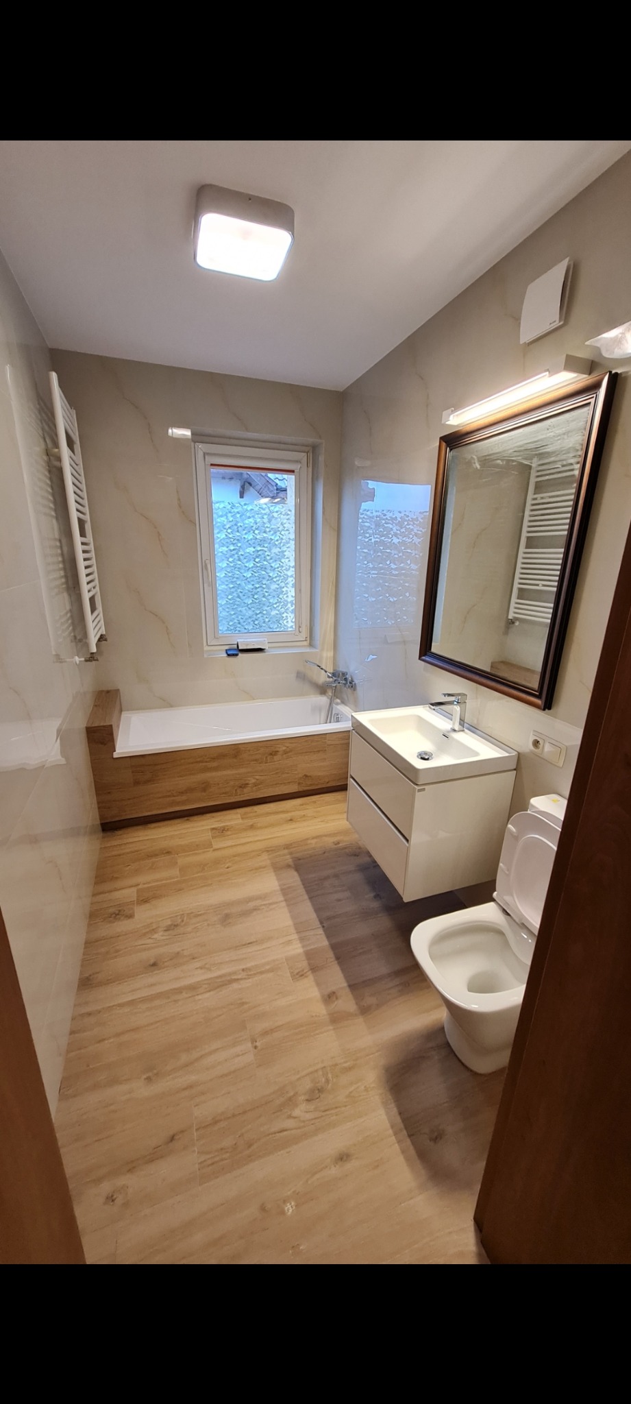 Jasna łazienka po remoncie z drewnianą obudową wanny, beżowymi płytkami imitującymi marmur i drewnopodobną podłogą, widok na umywalkę z szafką i toaletę.