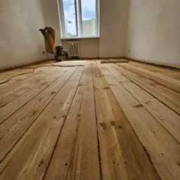 Cyklinowanie drewnianej podłogi z widoczną maszyną do cyklinowania i poziomnicą w jasnym, pustym pomieszczeniu z oknem.