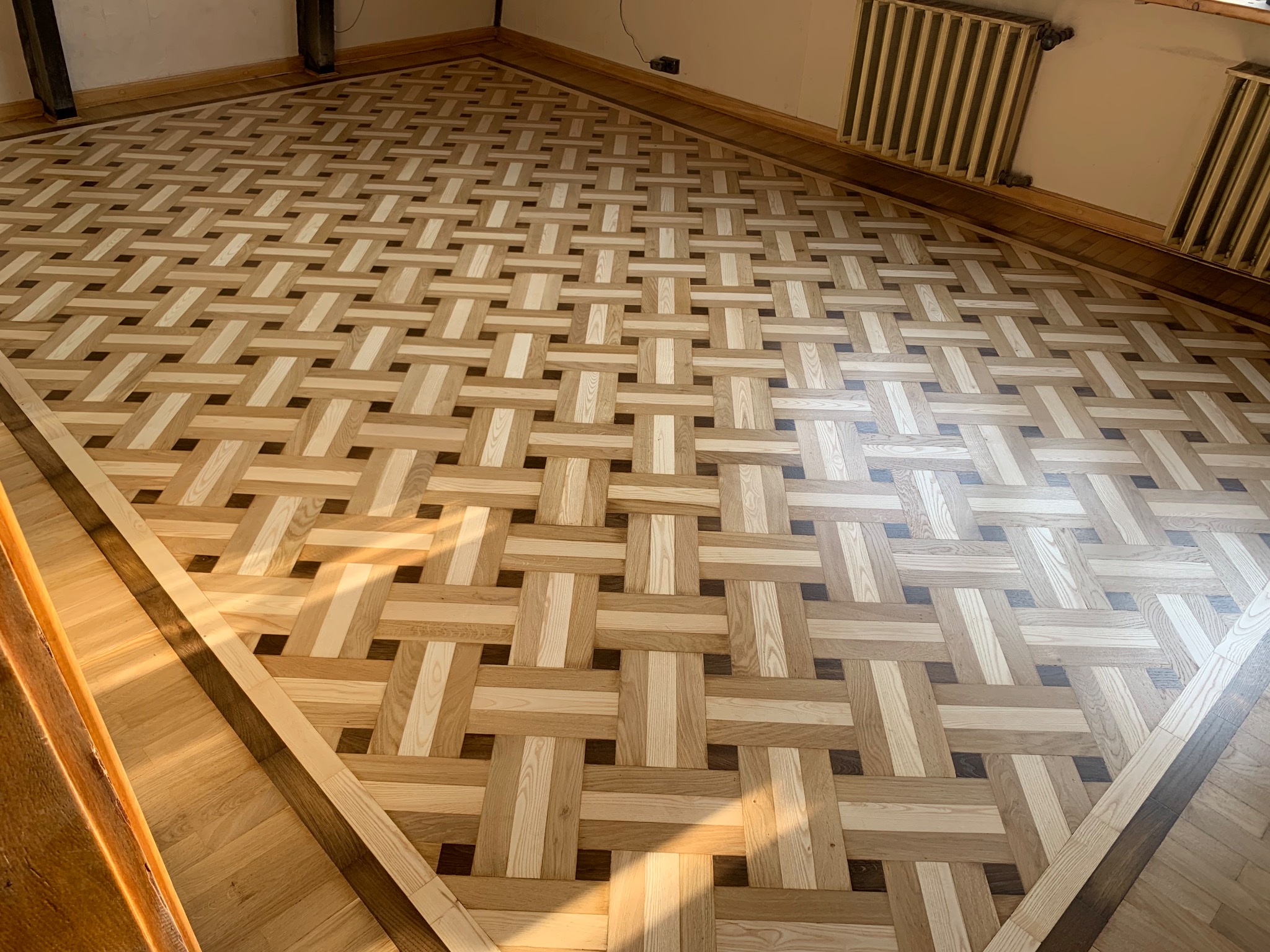 Wzór parkietu ułożonego w jodełkę koszykową z jasnego i ciemniejszego drewna, widoczne grzejniki żeliwne przy ścianie.