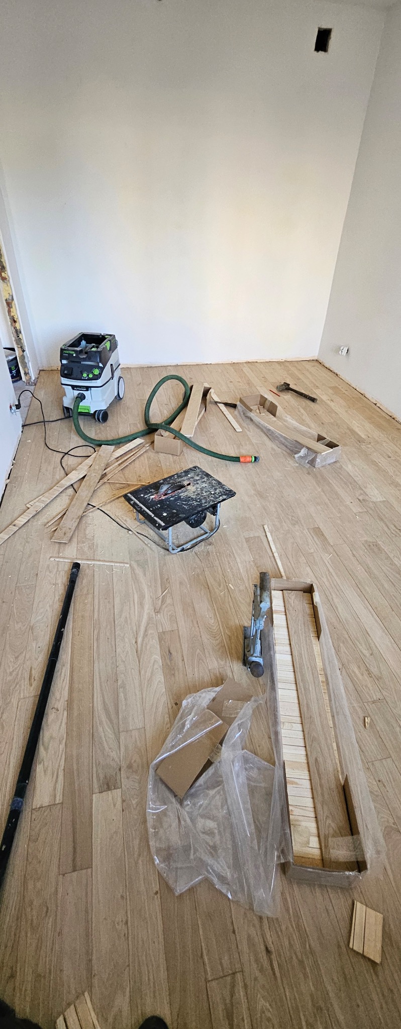 Fragment pokoju w trakcie montażu drewnianej podłogi, widoczne narzędzia: odkurzacz przemysłowy, piła stołowa, dobijak pneumatyczny, oraz resztki materiału i opakowania.
