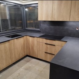Łukasz Suchocki - Nowoczesna kuchnia z drewnianymi frontami i czarnym blatem, minimalistyczne uchwyty, okno nad zlewem, płyta indukcyjna. Stonowana kolorystyka, elegancki design.