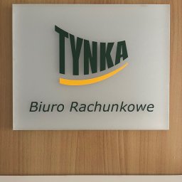 BIURO RACHUNKOWE TYNKA JUSTYNA WARWAS - Szyld z nazwą 'TYNKA Biuro Rachunkowe' na drewnianej ścianie, zielony napis z żółtym akcentem.