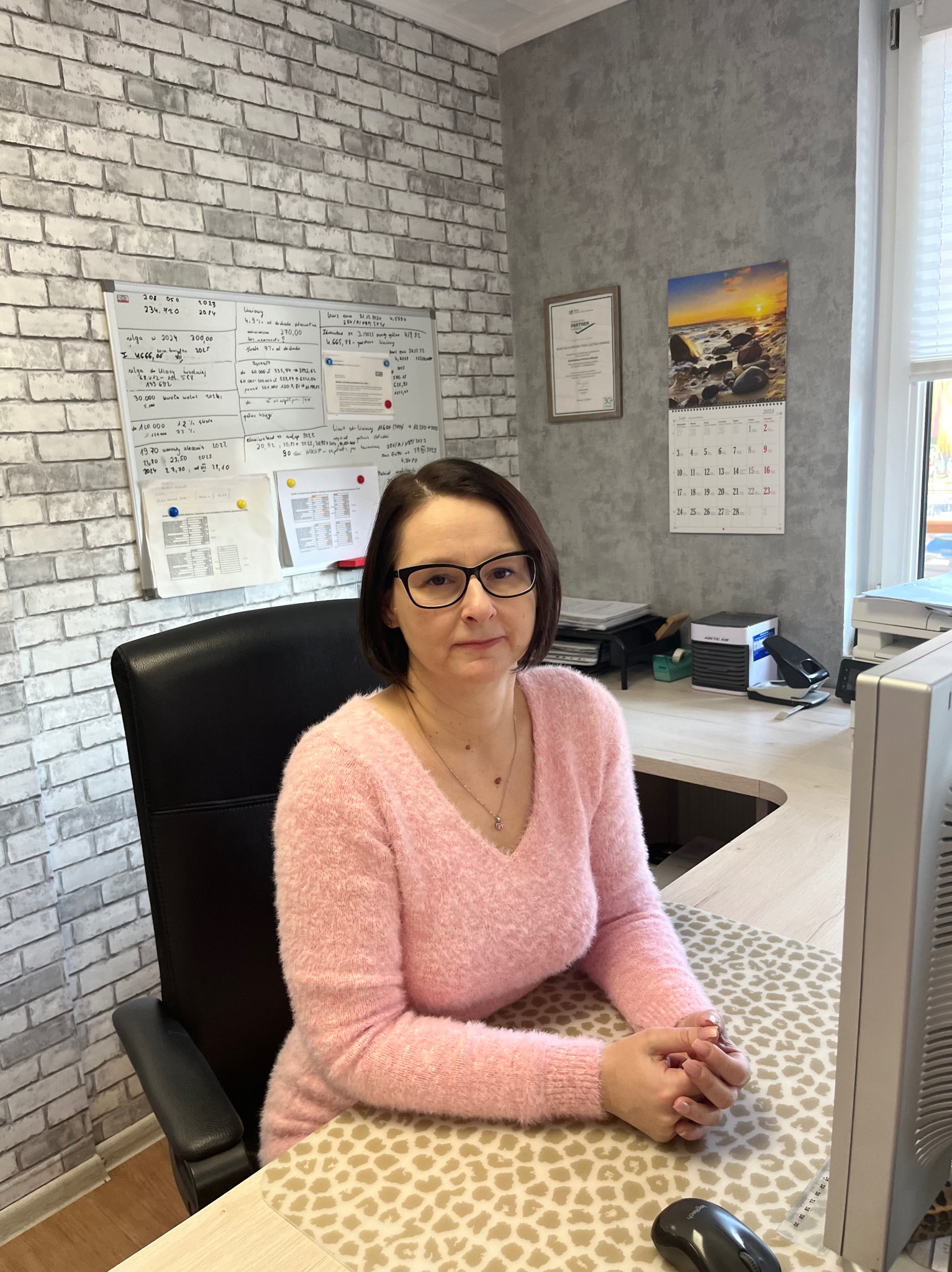 Kobieta w okularach siedzi przy biurku z blatem imitującym panterkę, za nią biała tablica z zapiskami, kalendarz i certyfikat na szarej ścianie z efektem betonu.