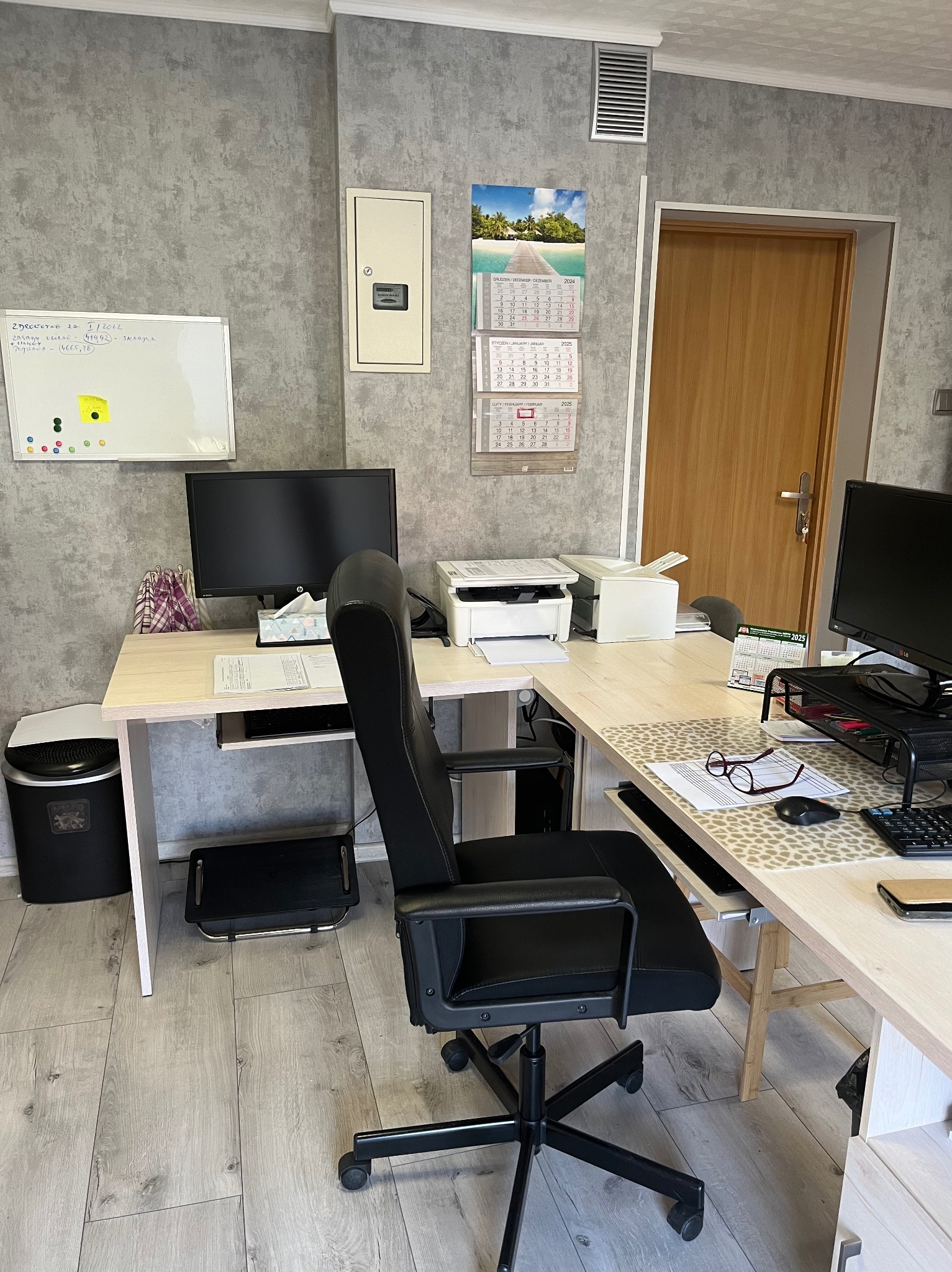 Biuro z dwoma biurkami w kształcie litery L, z komputerami, drukarkami, kalendarzem i tablicą suchościeralną, utrzymane w szarych i beżowych tonacjach.