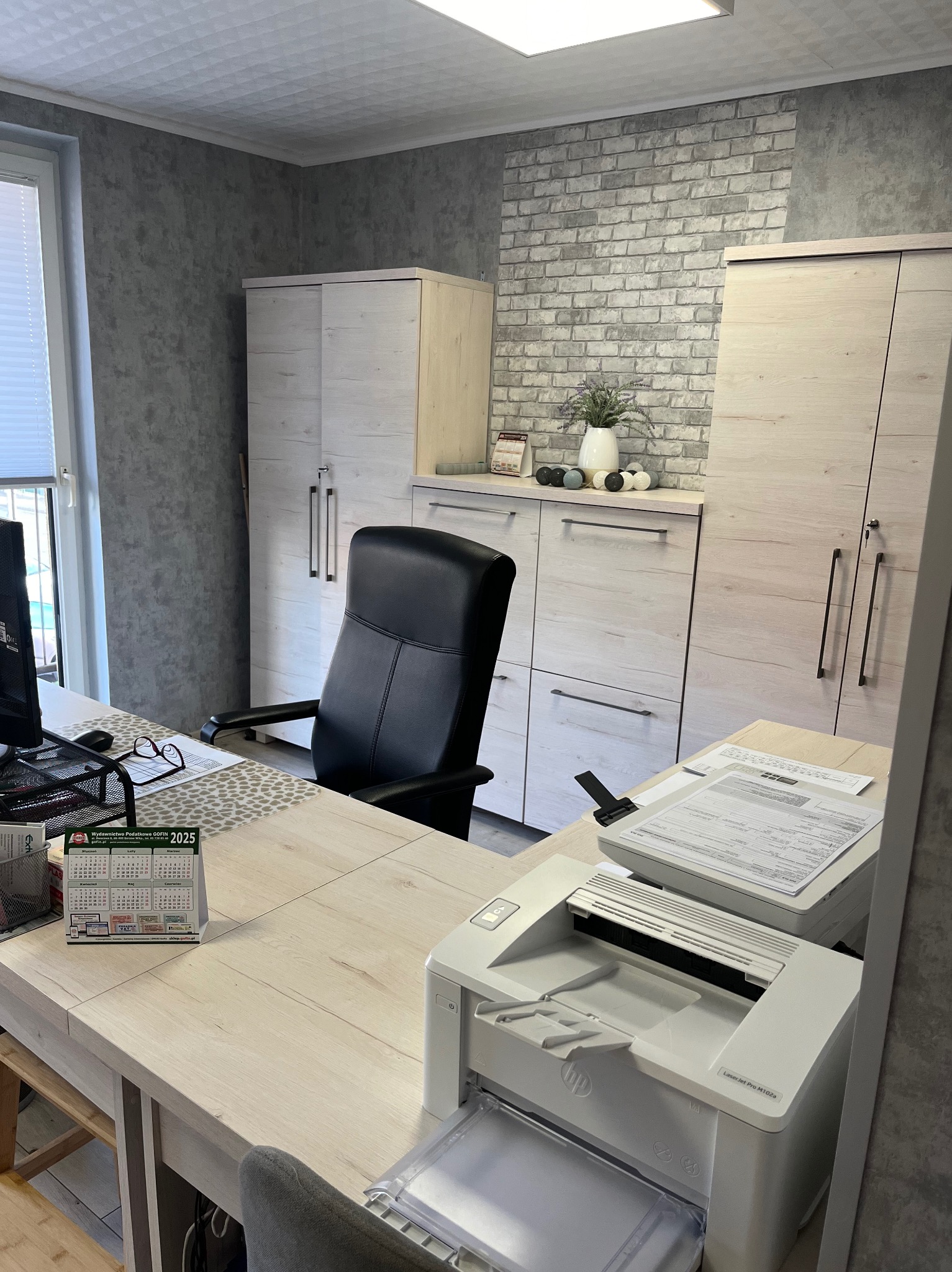 Biuro z biurkiem, krzesłem, szafkami i drukarką HP LaserJet Pro M102a, kalendarz Wydawnictwo Podatkowe GOFIN 2025 na biurku.