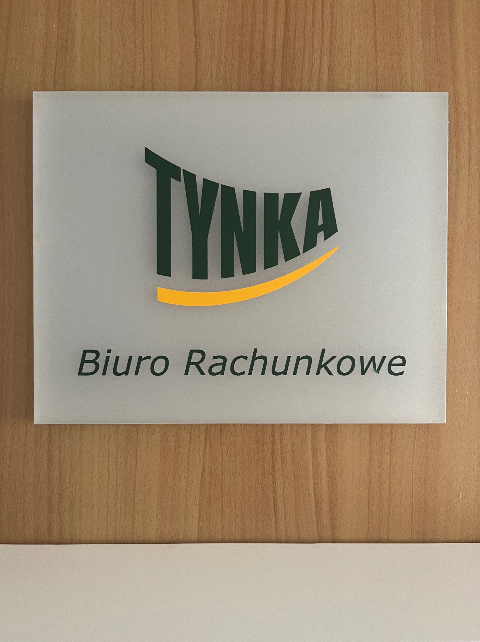 Szyld z nazwą 'TYNKA Biuro Rachunkowe' na drewnianej ścianie, zielony napis z żółtym akcentem.