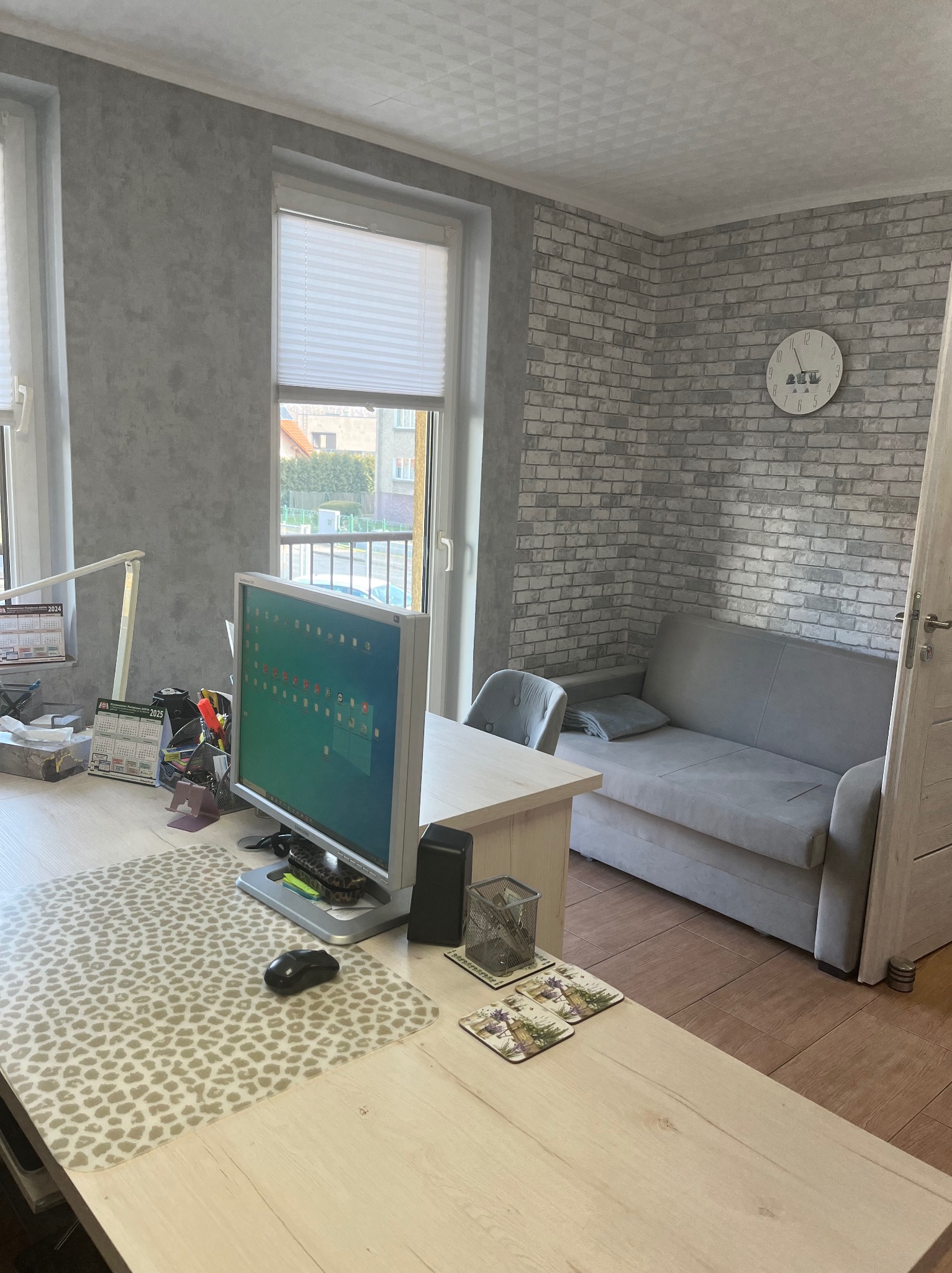 Biuro z komputerem, kalendarzami i akcesoriami biurowymi na biurku z podkładką w panterkę, w tle kanapa i ściana z cegły.