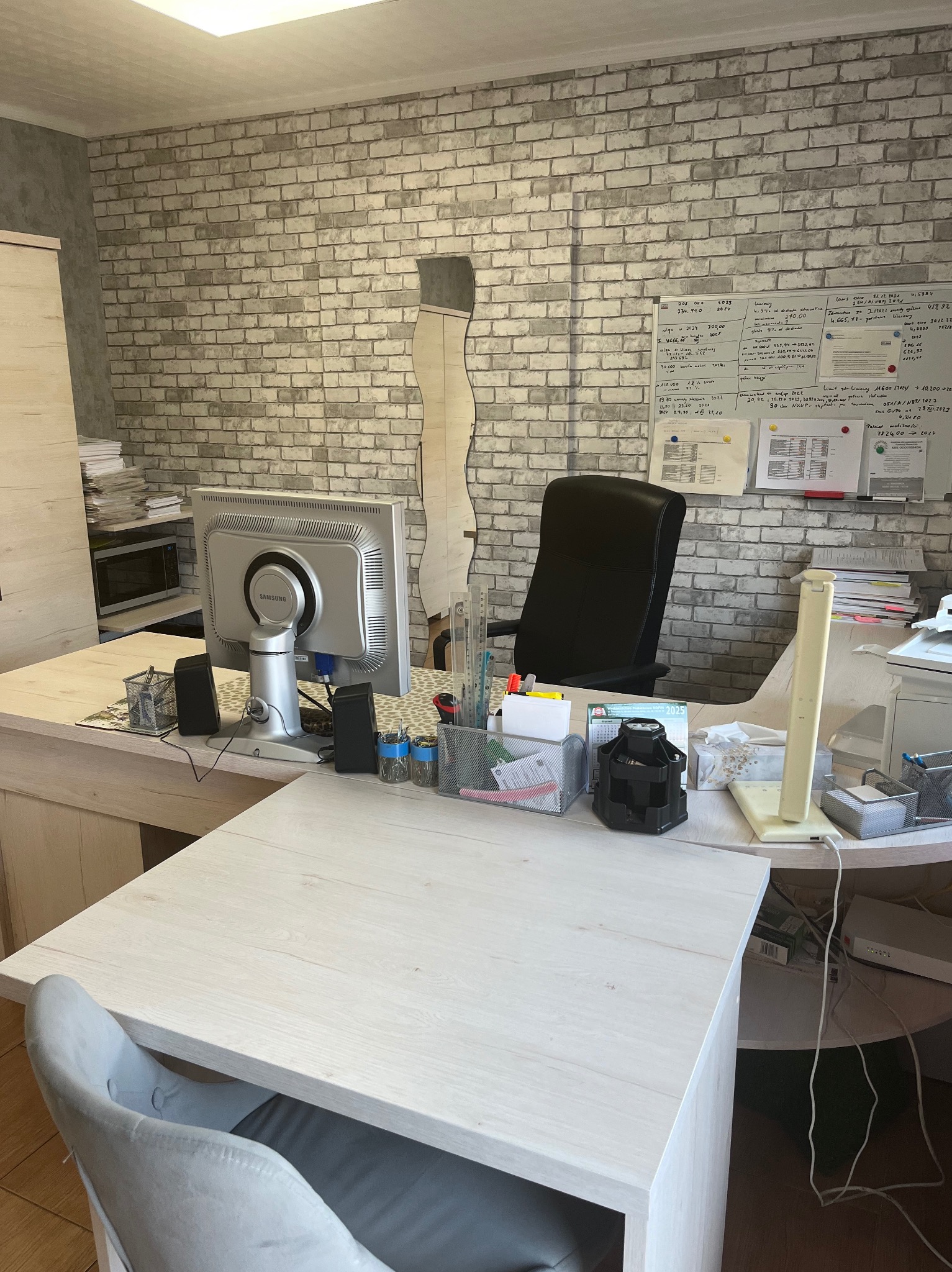 Biuro z biurkiem z jasnego drewna, krzesłem, monitorem Samsung, ścianą z cegły i tablicą z odręcznymi notatkami.