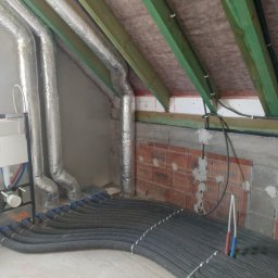 Modern House Solutions Damian Maziarz - Szare, elastyczne rury wentylacyjne ułożone rzędami na podłodze poddasza, z widocznymi elementami instalacji hydraulicznej i izolowanymi przewodami wentylacyjnymi przy ścianie.