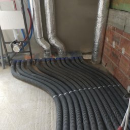 Modern House Solutions Damian Maziarz - Szereg czarnych, karbowanych rur wentylacyjnych podłączonych do rozdzielacza z niebieskimi końcówkami, z widocznymi izolowanymi przewodami oraz aluminiowymi kanałami wentylacyjnymi w surowym...