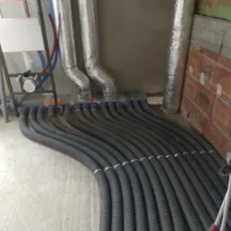 Szereg czarnych, karbowanych rur wentylacyjnych podłączonych do rozdzielacza z niebieskimi końcówkami, z widocznymi izolowanymi przewodami oraz aluminiowymi kanałami wentylacyjnymi w surowym...