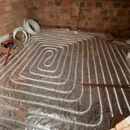 Instalacja ogrzewania podłogowego z białych rurek ułożonych w pętle na srebrnej folii z czerwoną siatką na tle ceglanej ściany i podłogi w trakcie montażu.