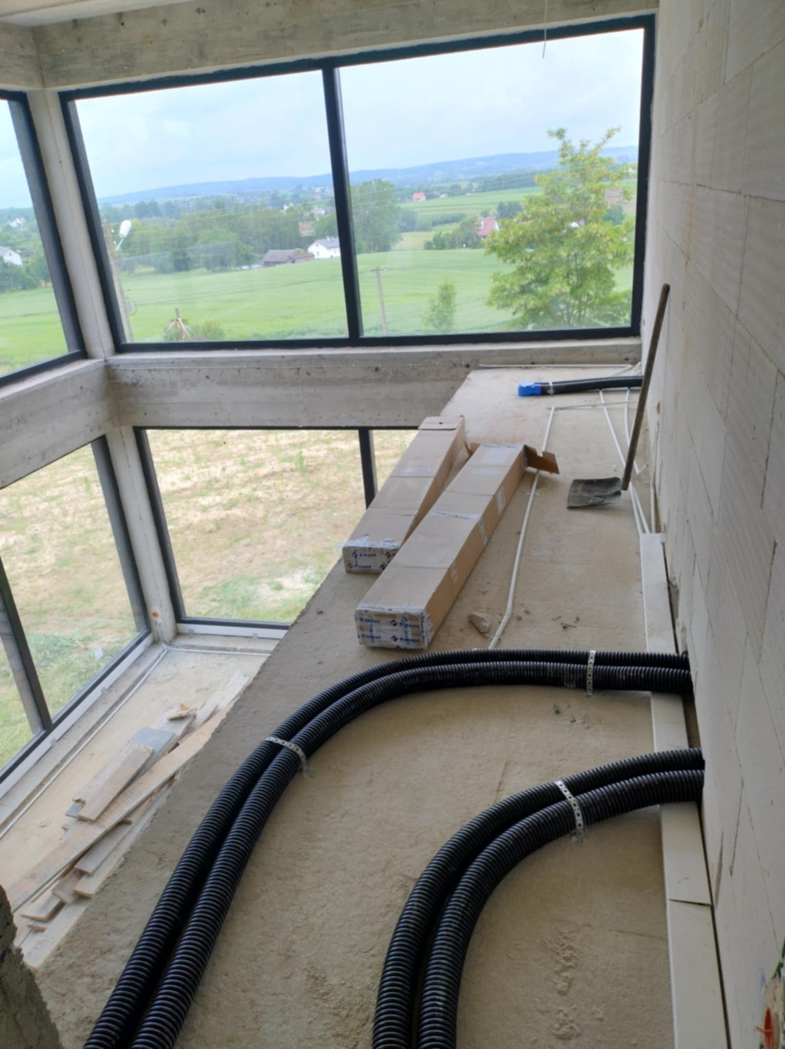 Surowy stan wnętrza z widokiem na krajobraz przez duże okna; widoczne czarne rury wentylacyjne przymocowane do ściany, kartony z elementami instalacji, oraz narzędzia do montażu.