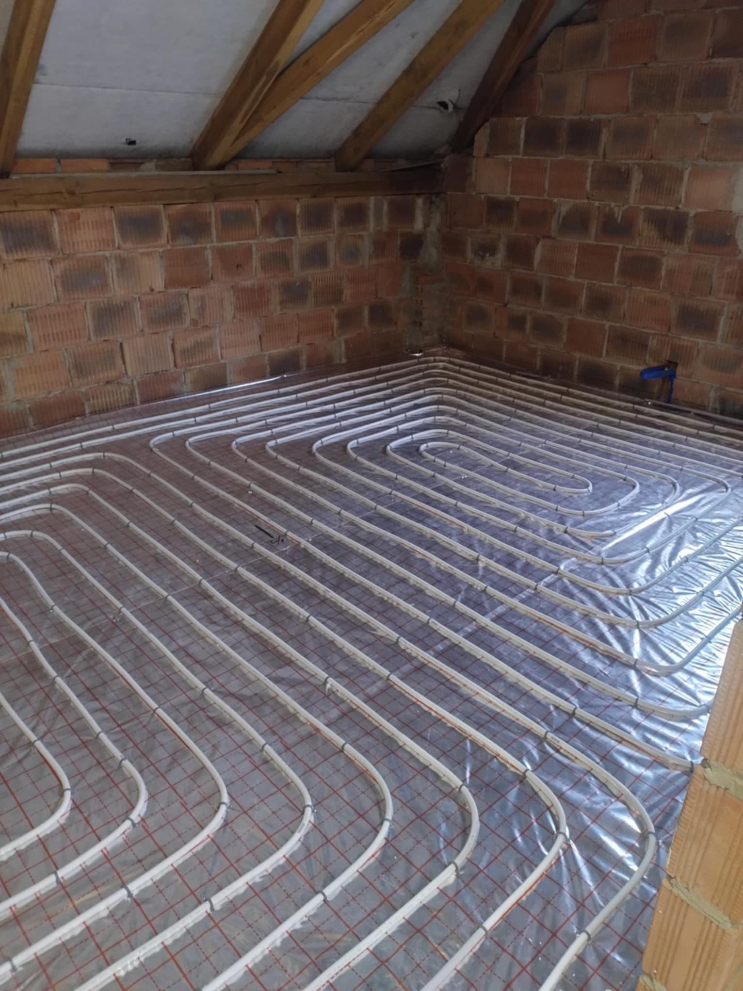 Białe rury ogrzewania podłogowego ułożone spiralnie na folii z czerwoną siatką w surowym wnętrzu z ceglanymi ścianami i drewnianą konstrukcją dachu.