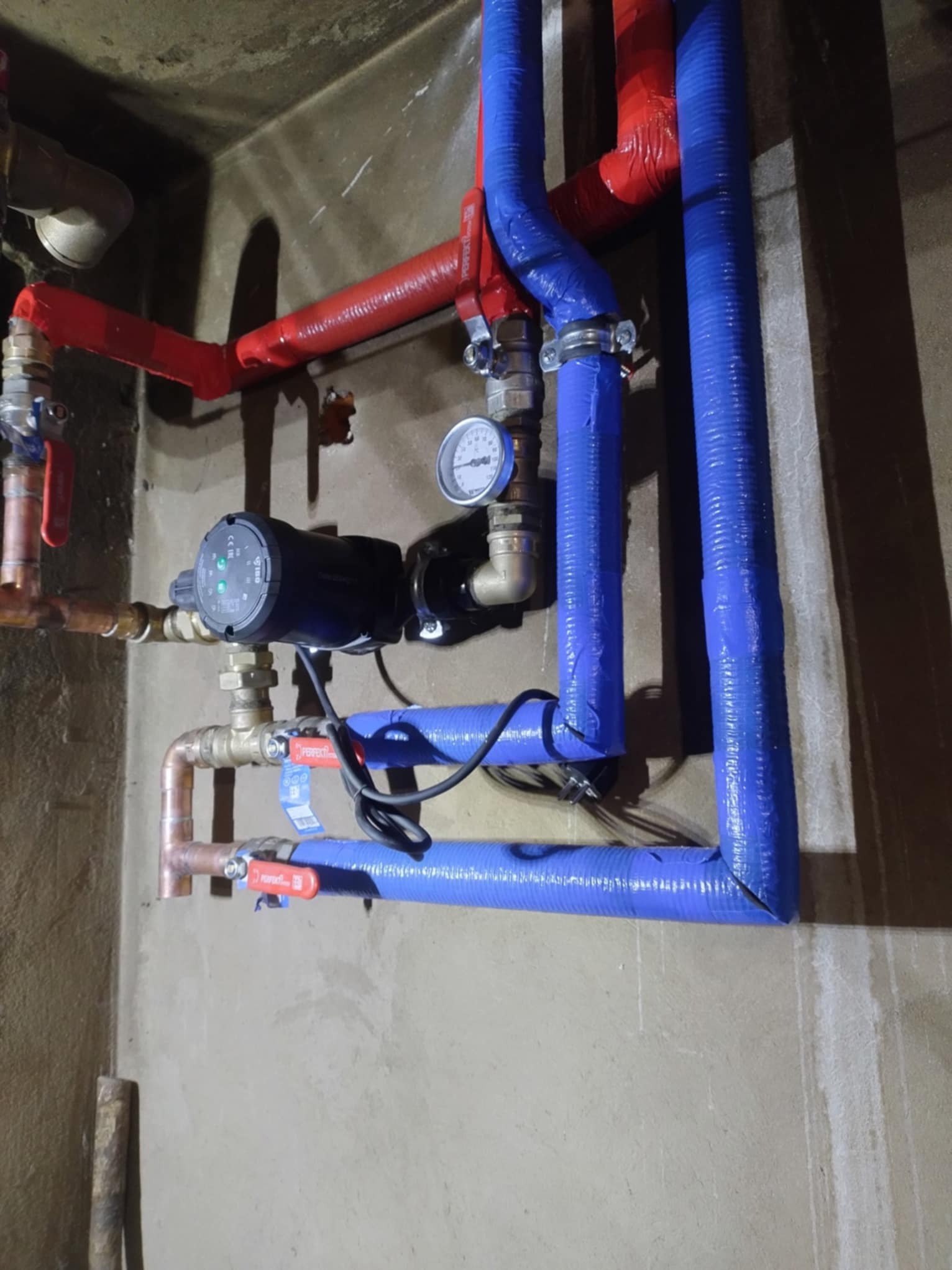 Instalacja hydrauliczna z pompą obiegową, rurami w izolacji termicznej w kolorach czerwonym i niebieskim, manometrem i zaworami odcinającymi, zamontowana na surowej ścianie.