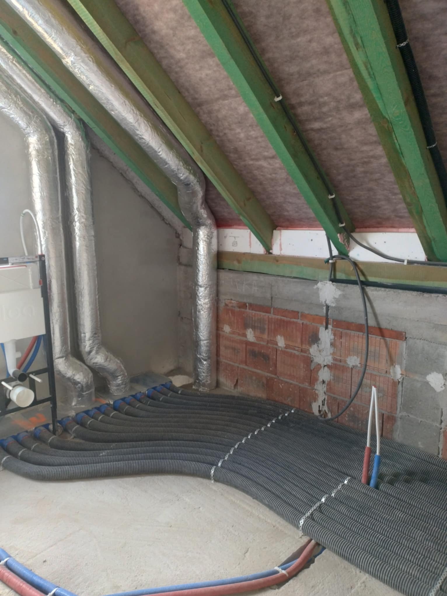 Szare, elastyczne rury wentylacyjne ułożone rzędami na podłodze poddasza, z widocznymi elementami instalacji hydraulicznej i izolowanymi przewodami wentylacyjnymi przy ścianie.
